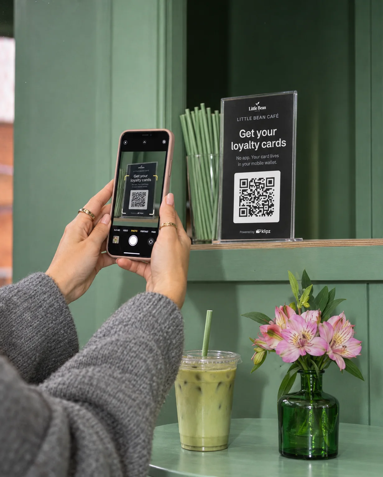 In a sage-green café, a customer in a grey jumper holds up her phone with both hands and scans the QR stand on the hatch's ledge, her iced matcha waiting on the table beside a vase of pink flowers.